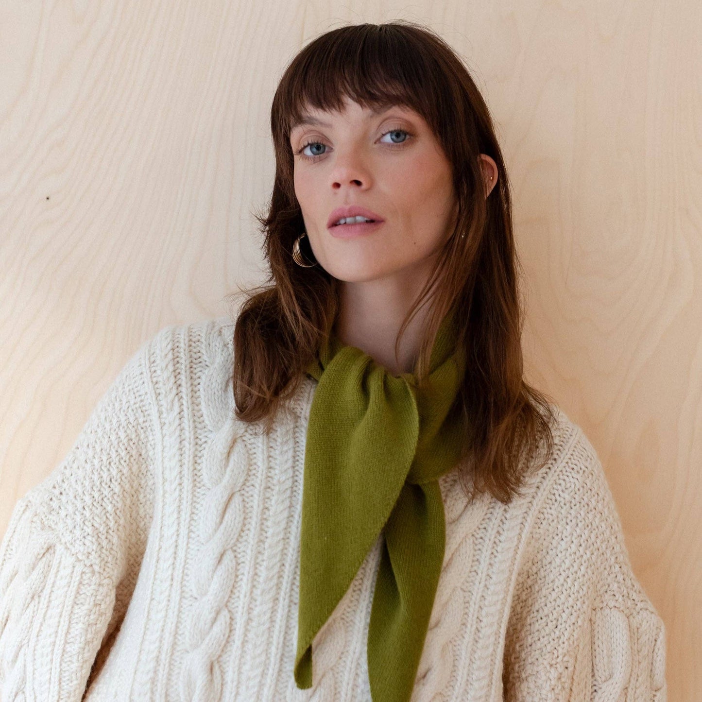 Woman wearing a white cable knit sweater with a green scarf against a beige background