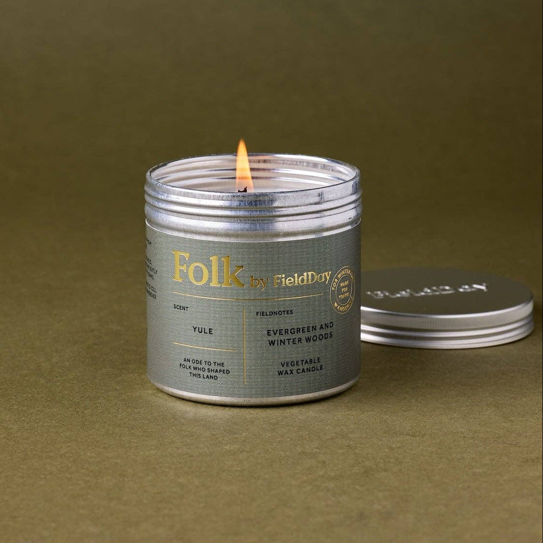 A lit silver metal candle tin with a white label reading 'Folk by FieldDay' sits on a textured olive green surface, with its lid resting beside it.