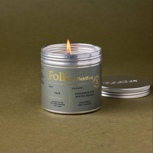 A lit silver metal candle tin with a white label reading 'Folk by FieldDay' sits on a textured olive green surface, with its lid resting beside it.